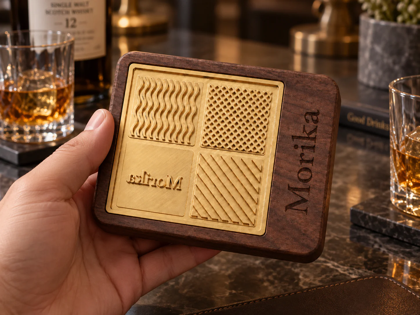 Hand holding a copper and walnut ice branding stamp on a bar beside whiskey glasses